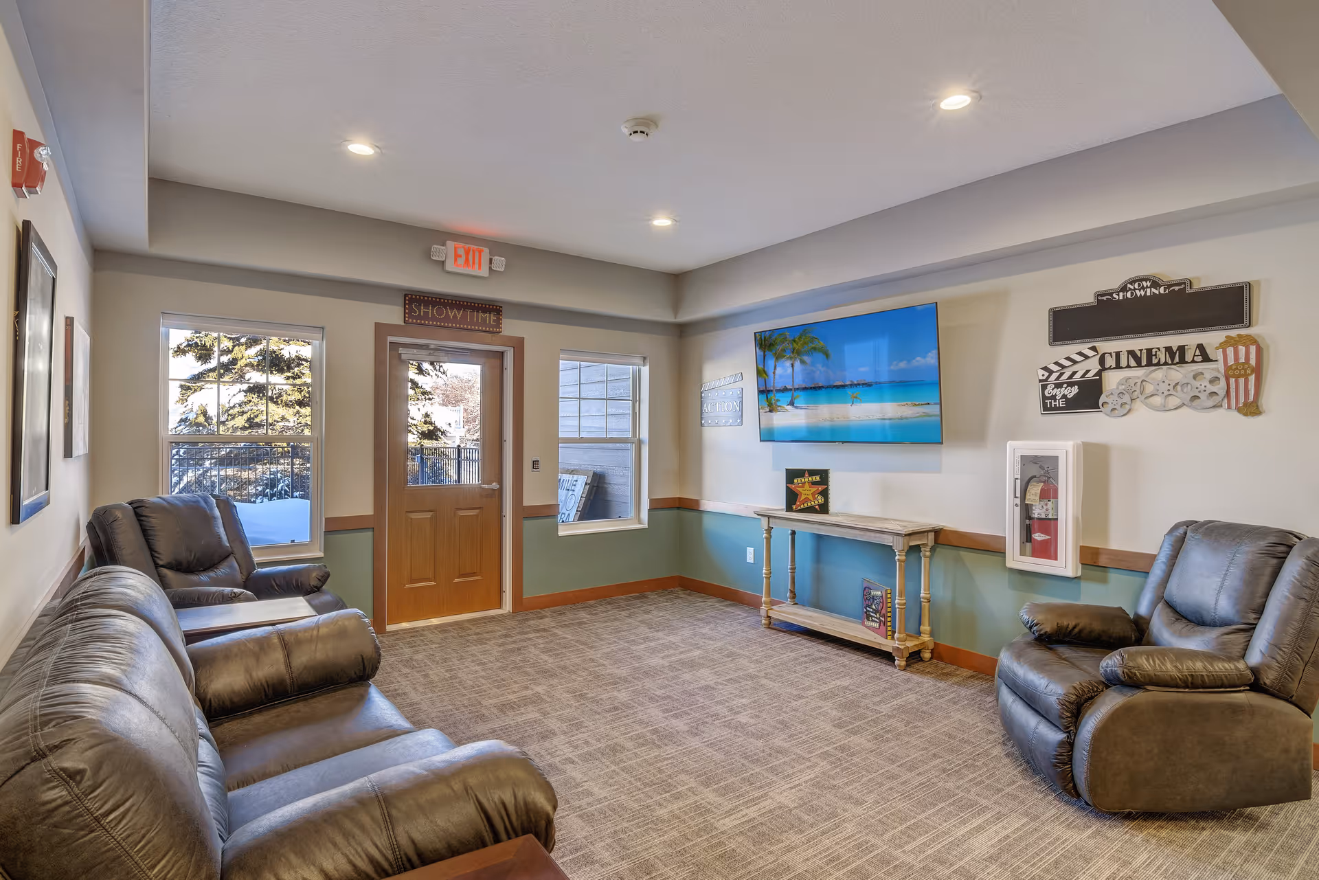 A cozy living room area in a senior living facility with leather sofas and recliners arranged around a wall-mounted flat-screen TV displaying a beach scene. The room has beige walls with a green lower half, carpeted floor, and decorative cinema-themed wall art. There are two windows and a wooden door with an exit sign above it.