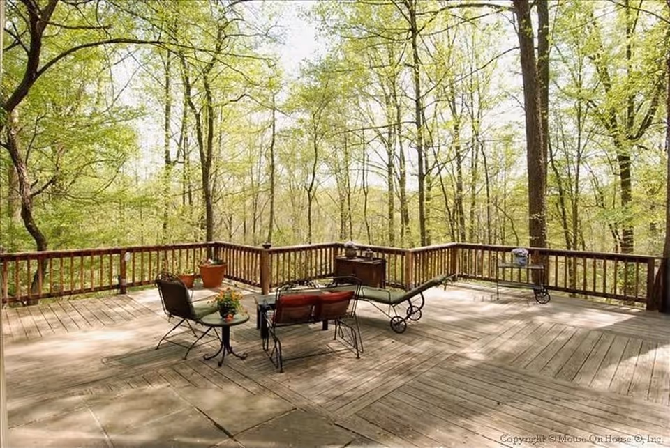 A spacious wooden outdoor deck surrounded by trees with fresh green leaves. The deck has a wooden railing and is furnished with a small round table, two chairs, a cushioned bench, a lounge chair, and a small cart. There are potted plants on the deck, and sunlight filters through the trees creating a peaceful natural setting.