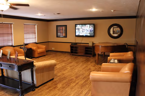 A cozy living room with wooden flooring, beige walls, and brown trim. The room features a flat-screen TV mounted on the wall, a wooden console table beneath it, a grand piano, and several comfortable leather and upholstered armchairs arranged around the space. There are two windows with wooden blinds and framed artwork on the walls.