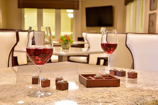 A close-up view of a granite table with two glasses of red wine and a wooden tic-tac-toe game set. In the background, there are cream-colored upholstered chairs and a vase with flowers on another table in a well-lit room.