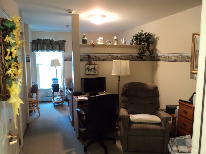 Interior view of a senior living facility room at Updyke's Willow Ridge showing a cozy seating area with a cushioned recliner chair and a floor lamp beside it. A desk with a computer monitor and an office chair is positioned against a partial wall. Decorative items and plants are placed on a shelf above the desk. In the background, there is a window with curtains, a standing lamp, and wooden chairs. The walls have a decorative border and the room is softly lit.