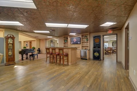 Open interior common area with a small bar and stools, a piano and seating, and a view into a dining room.
