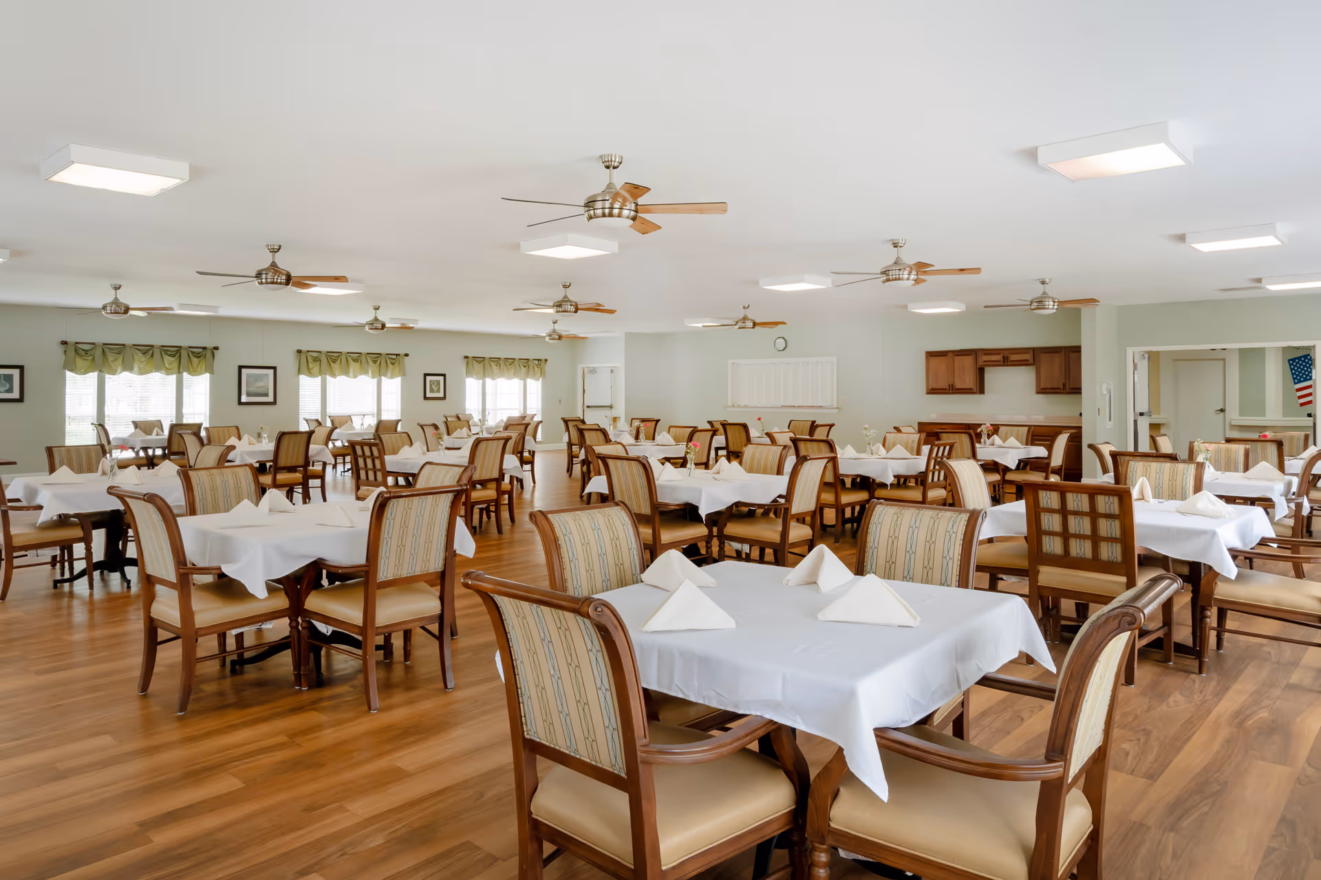 Spacious senior living dining room with multiple tables set with white tablecloths and folded napkins, wooden chairs, ceiling fans, and large windows.