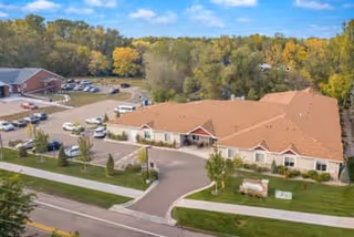 Aerial view of a single-story senior living facility with a large parking lot, surrounded by trees with autumn foliage under a blue sky with scattered clouds.
