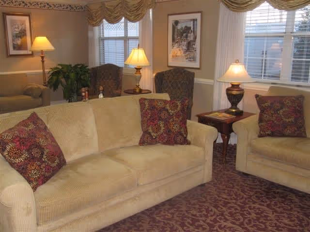 Cozy living room with beige sofas and armchairs, patterned throw pillows, table lamps, and windows with valances.