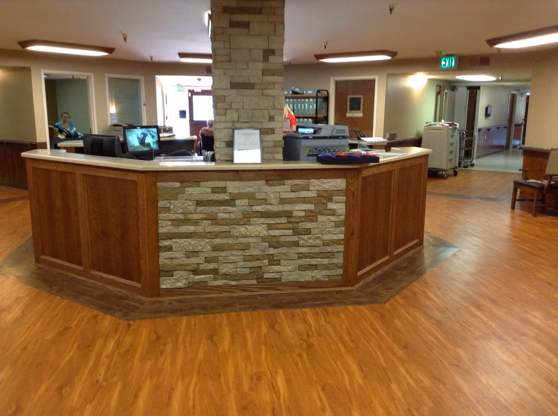Interior view of a senior living facility reception area with a wooden and stone front desk in the center. There are computer monitors, office supplies, and staff members behind the desk. The floor is wood laminate, and there are hallways leading to other rooms in the background.