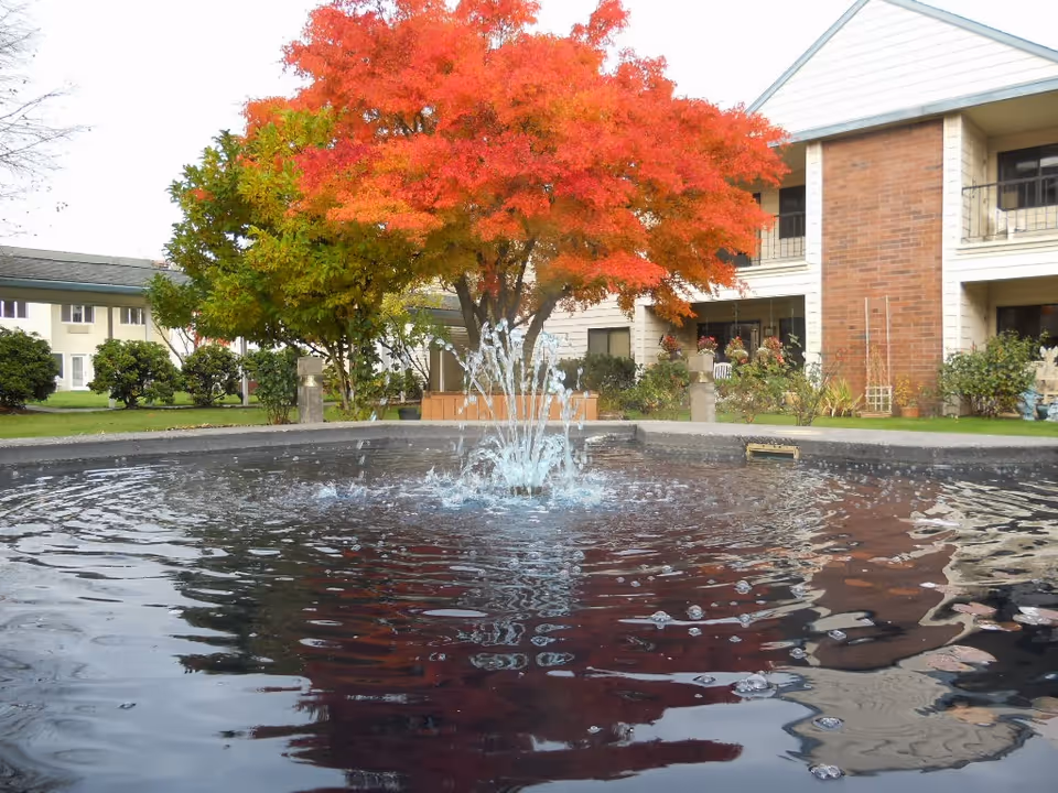 A courtyard area with a water fountain in the center, surrounded by a circular basin. Behind the fountain, there is a tree with vibrant red and orange autumn leaves, along with other green bushes and plants. In the background, there is a two-story building with balconies and windows, part of the Jennings McCall Center facility.
