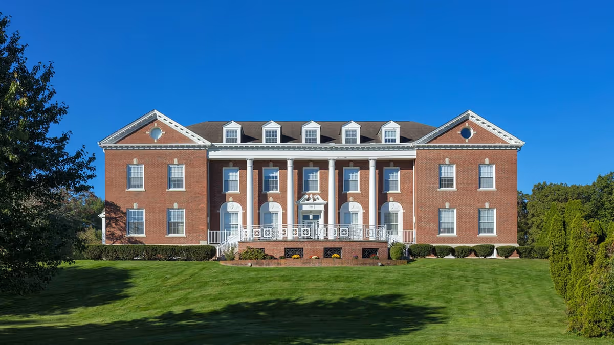 Front exterior view of a large, stately brick building with white columns and multiple windows, surrounded by a well-maintained green lawn and trees under a clear blue sky.