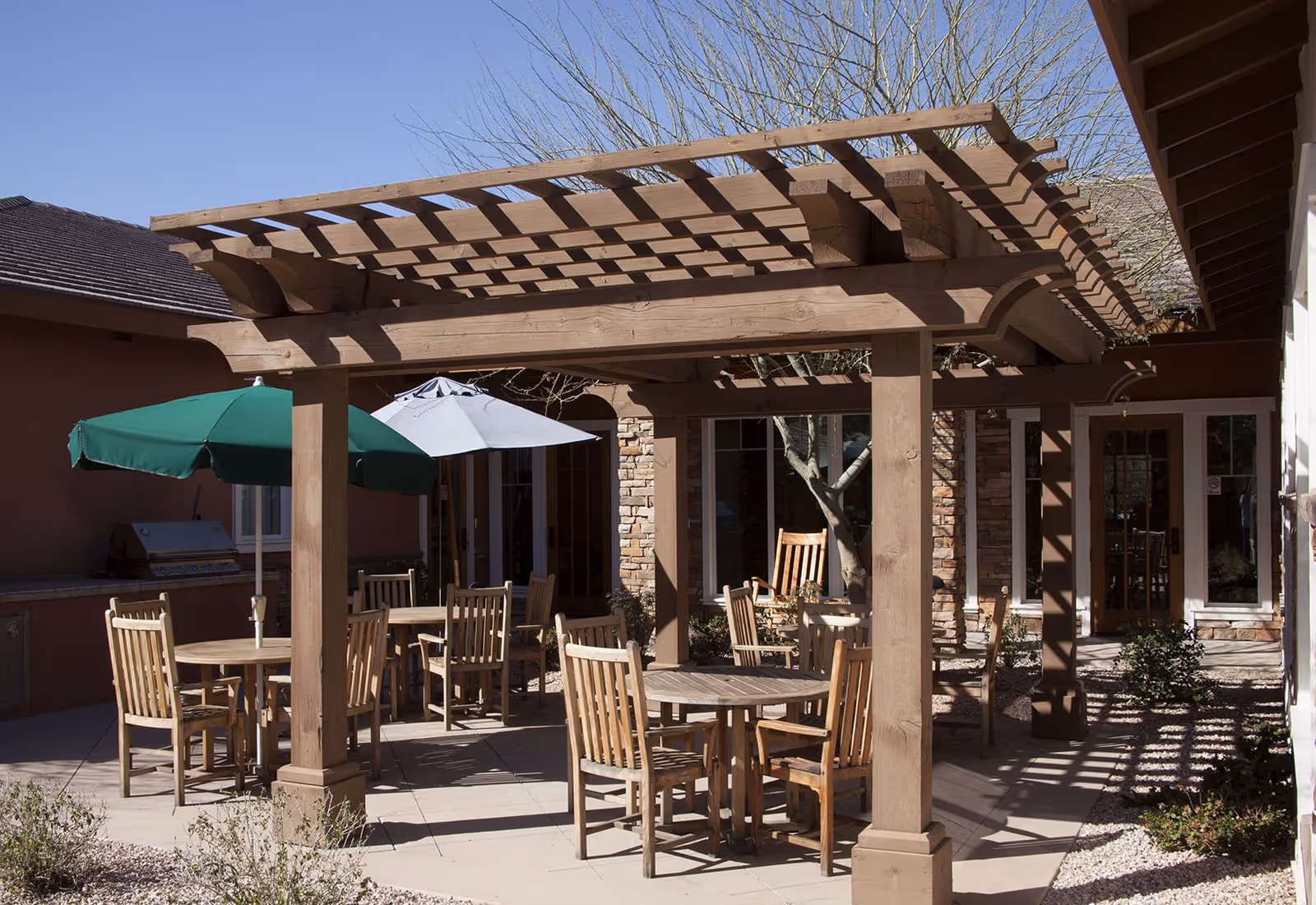 Outdoor patio area with wooden pergola providing partial shade over round wooden tables and chairs. Two umbrellas, one green and one white, are open over some tables. The patio is surrounded by buildings with stone and stucco walls, and there are some small plants and trees around the area.