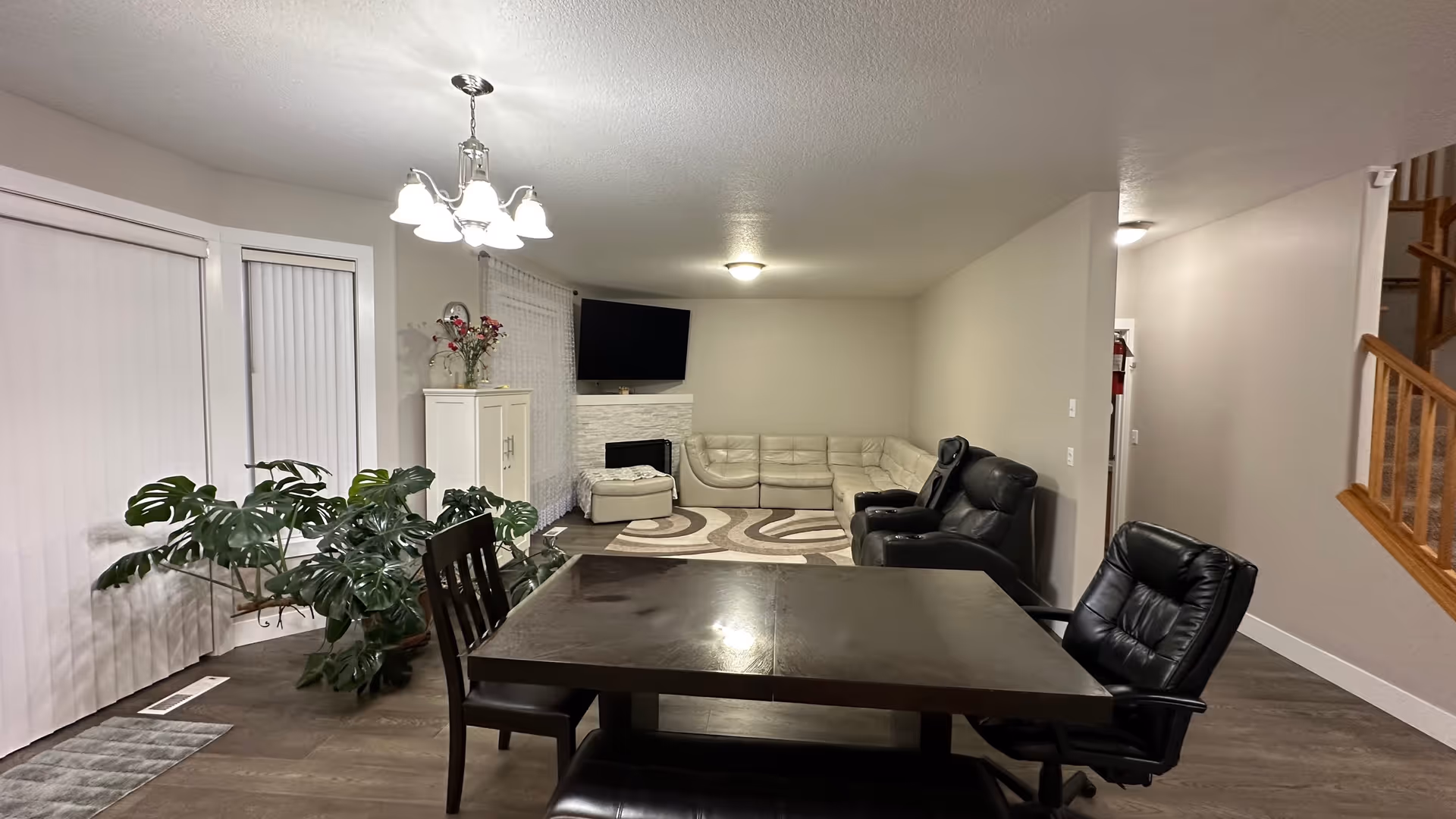 Interior view of a living and dining area with a dark wooden dining table and chairs in the foreground, a large green plant near sliding glass doors with vertical blinds, a white cabinet with flowers on top, a wall-mounted TV above a white fireplace, a beige sectional sofa, two black recliner chairs, and a staircase with wooden railing on the right side.