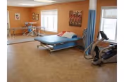 A rehabilitation room with a blue padded therapy table, parallel bars for walking exercises, an exercise bike, and a window with white blinds. The walls are painted orange and decorated with framed pictures.