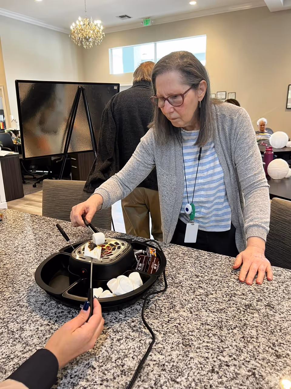 An elderly woman with glasses roasting marshmallows over a small indoor electric fire pit on a granite countertop. Another person is holding a marshmallow on a skewer towards the fire. The setting appears to be a communal indoor area with chairs and tables, and a chandelier is visible in the background.
