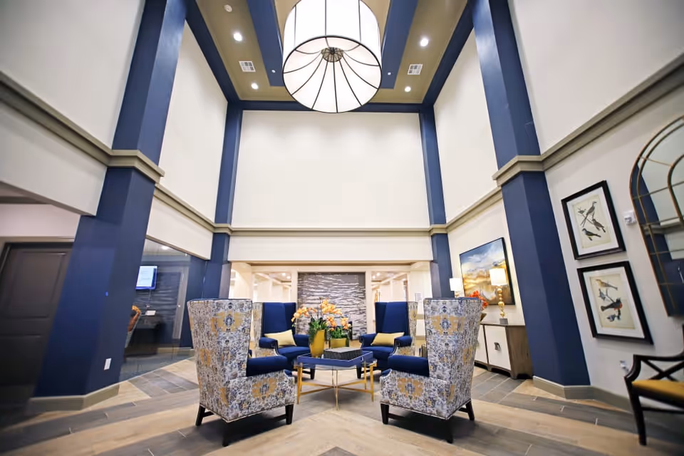 Bright double-height lobby with blue columns, patterned armchairs and blue sofas arranged around a central coffee table under a large hanging light fixture.