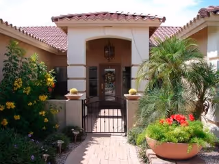 Front entrance of a residential building with a tiled roof, an arched doorway, a small black metal gate, and surrounding lush greenery including flowering plants and palm trees.