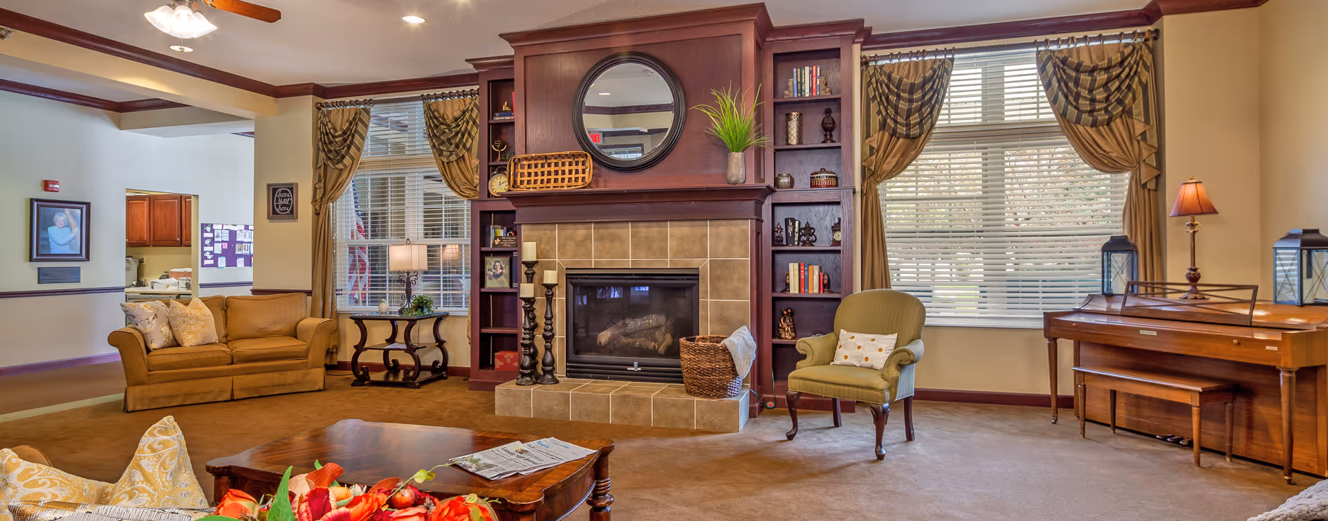 A cozy living room area in a senior living facility featuring a fireplace with a wooden mantel and built-in shelves, a round mirror above the fireplace, a green armchair with a pillow, a wooden coffee table with a newspaper and flowers, a sofa with cushions, large windows with striped curtains, a wooden piano with a lamp and lanterns on top, and warm lighting throughout the room.