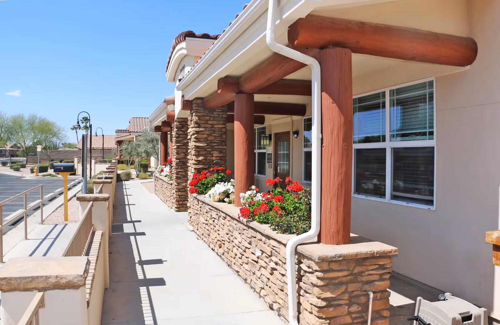 Exterior walkway of a senior living facility with stone pillars and wooden beams supporting a covered porch. Flower beds with red and white flowers line the walkway, and there are windows along the building wall. The sky is clear and blue.