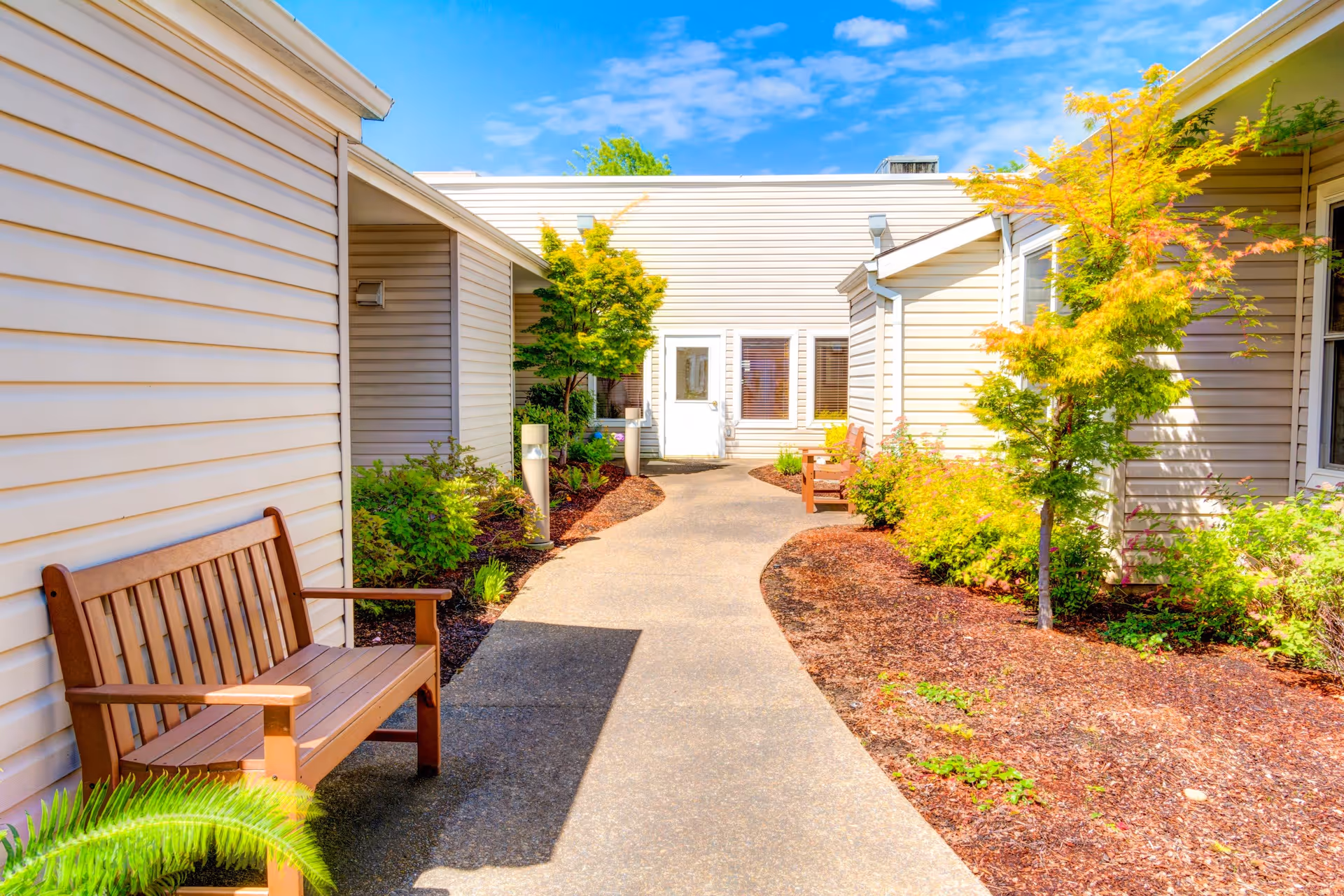 Outdoor walkway at Monterey Court Memory Care with beige siding buildings on both sides, wooden benches along the path, small trees, and shrubs under a bright blue sky.