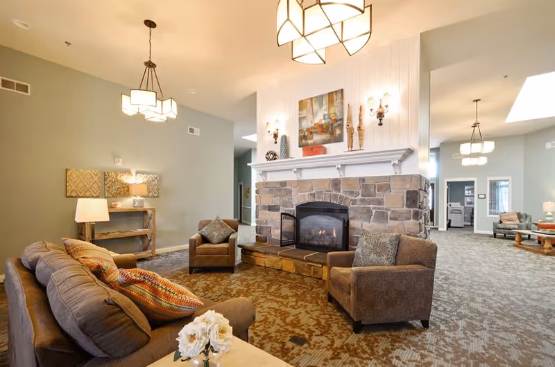 A cozy living room area with a stone fireplace in the center, flanked by two brown armchairs with patterned cushions. A brown sofa with colorful pillows faces the fireplace. The room has soft lighting from ceiling fixtures and wall sconces, with a decorative painting above the fireplace. The carpet has a patterned design, and there is a wooden shelf with a lamp and decor against a green wall.