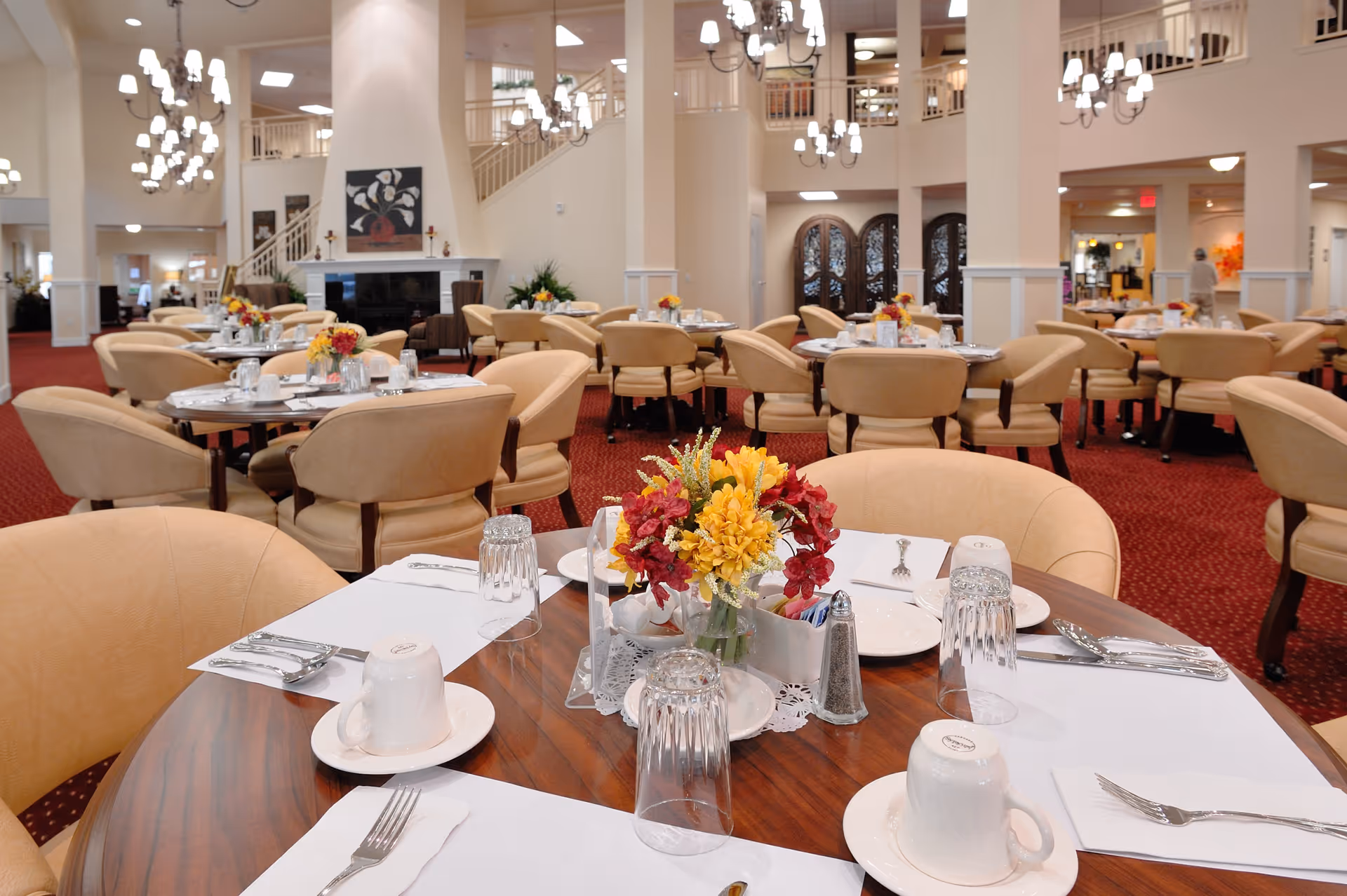 A spacious dining room with round tables set for a meal, featuring beige upholstered chairs, white placemats, cups, plates, silverware, and floral centerpieces. The room has red carpet flooring, cream-colored walls, chandeliers, and a fireplace with artwork above it. There are multiple tables and chairs arranged throughout the room, with a staircase and decorative doors visible in the background.
