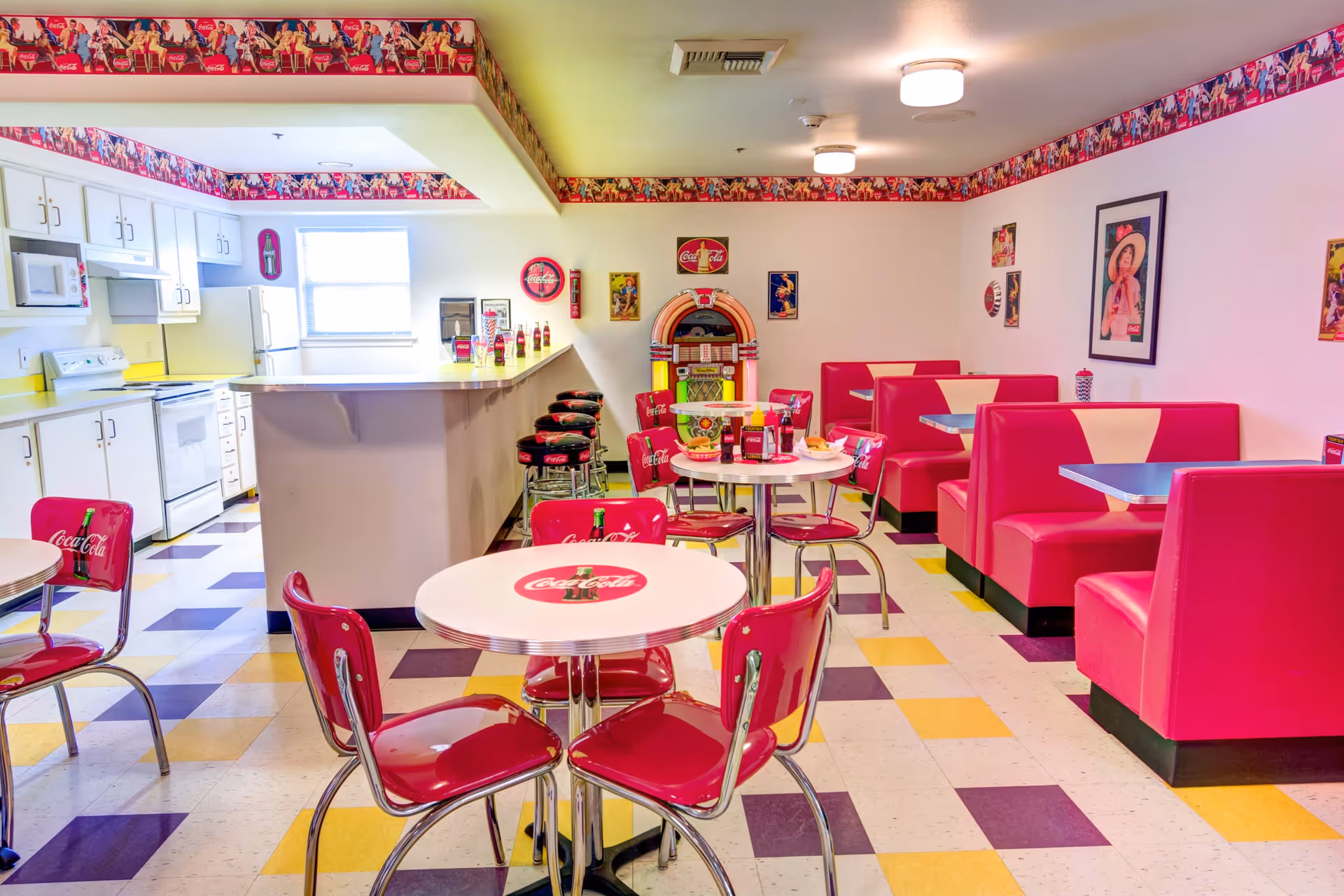 A retro-themed dining area with red Coca-Cola branded chairs and booths, round tables with Coca-Cola logos, a jukebox against the wall, and a kitchen area with white cabinets and appliances in the background. The floor has a checkered pattern with yellow, white, and purple tiles, and the walls are decorated with vintage Coca-Cola posters and a Coca-Cola border near the ceiling.