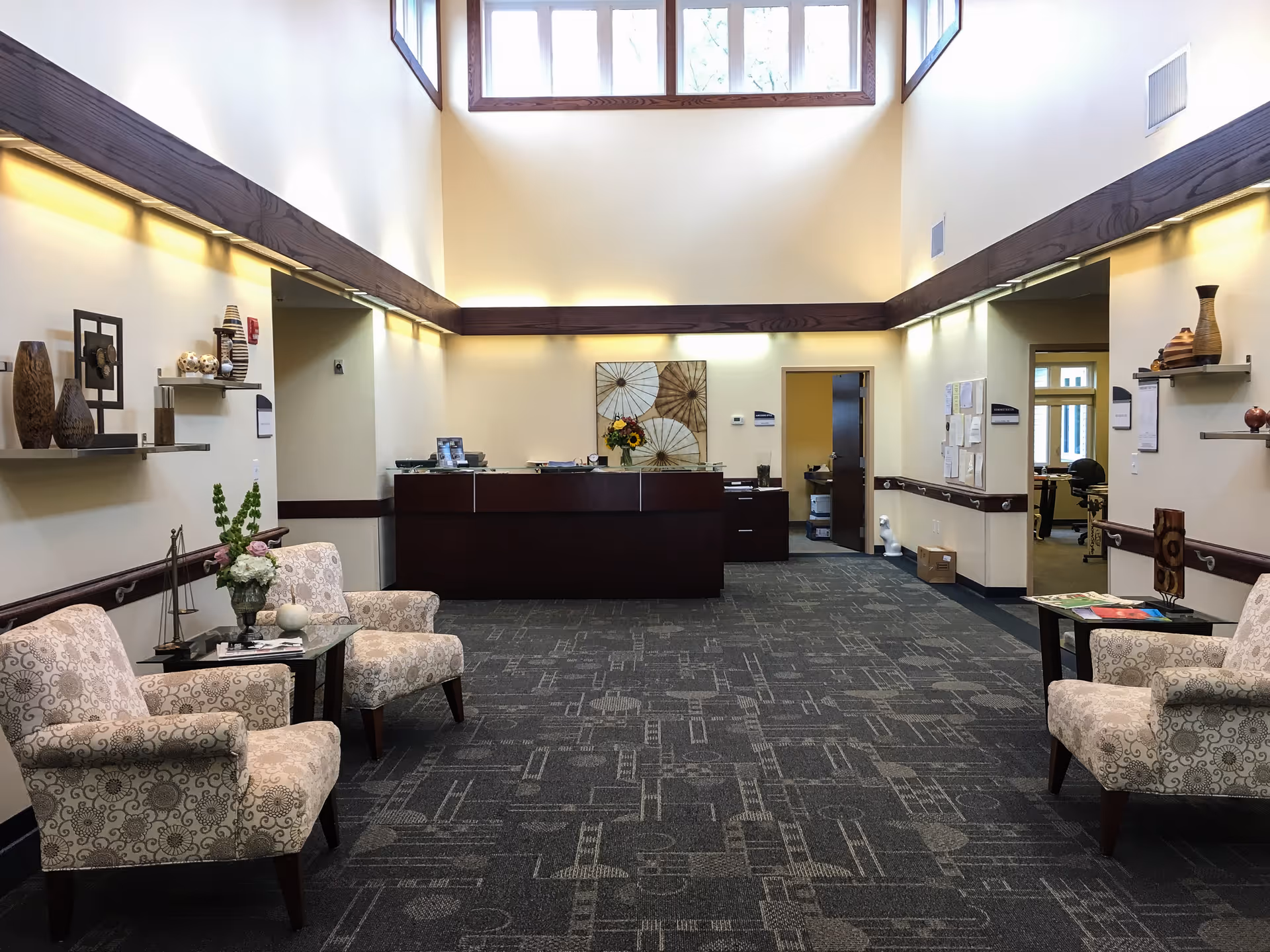 A spacious and well-lit reception area with high ceilings and large windows near the ceiling. The room features a dark wood reception desk at the far end, two patterned armchairs with a small glass table between them on each side of the room, and decorative shelves with vases and art pieces on the walls. The carpet has a geometric pattern, and there are doorways leading to other rooms on either side of the reception desk.