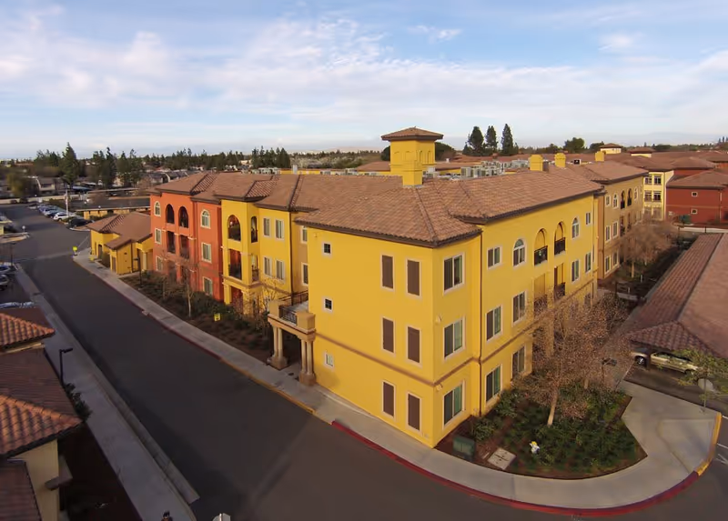 Aerial view of a three-story yellow and red Mediterranean-style apartment building with tiled roofs and surrounding road.