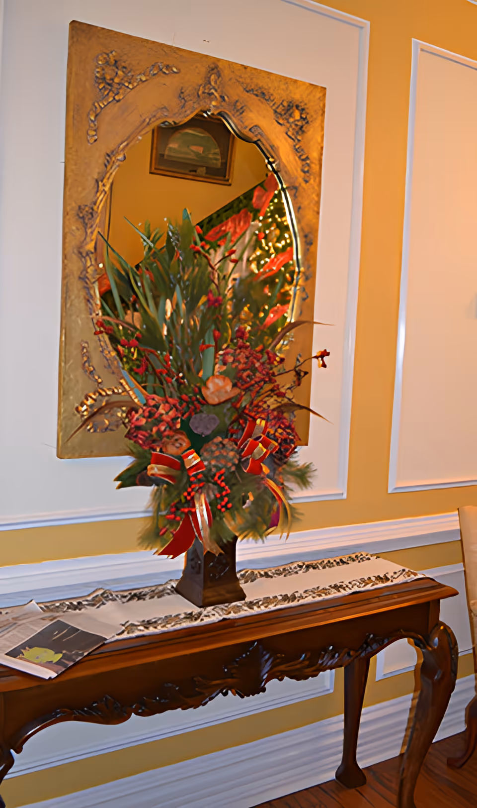 Ornate mirror above a wooden console table topped with a tall floral arrangement and decorative runner against a yellow wall.