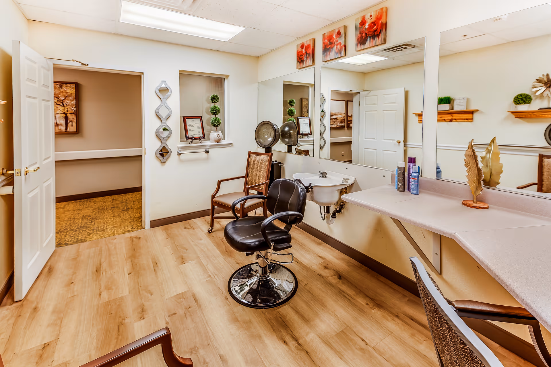Interior view of a salon or beauty room in a senior living facility with a black salon chair, a wooden chair, a sink, large mirrors on the wall, and shelves with decorative items and hair products. The room has light wood flooring and white walls with some artwork and plants.