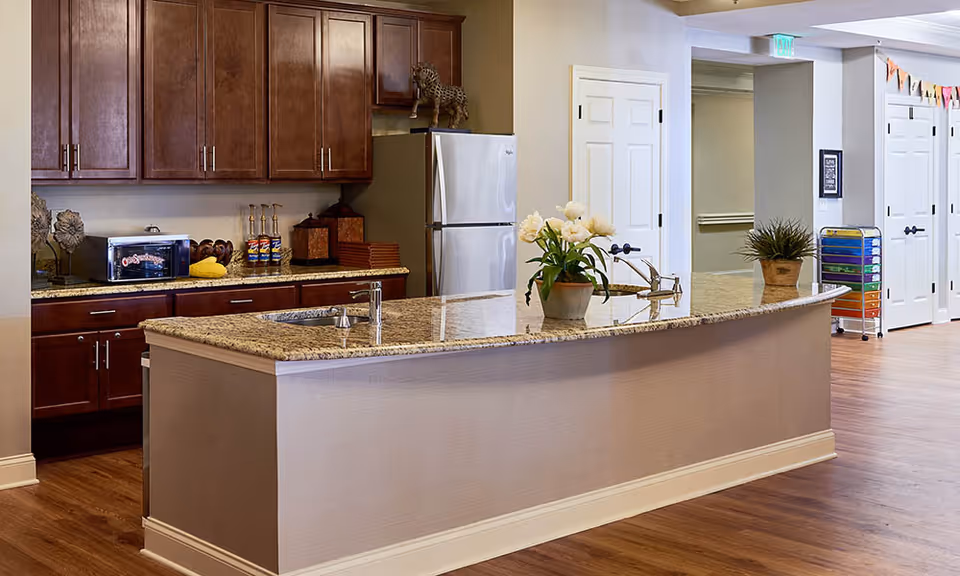 A modern kitchen area with a curved granite countertop island featuring two sinks and a potted plant with white flowers. Behind the island, there are dark wooden cabinets, a stainless steel refrigerator, a toaster oven, and various kitchen items on the countertop. The floor is wooden, and there are white doors and a hallway visible in the background.