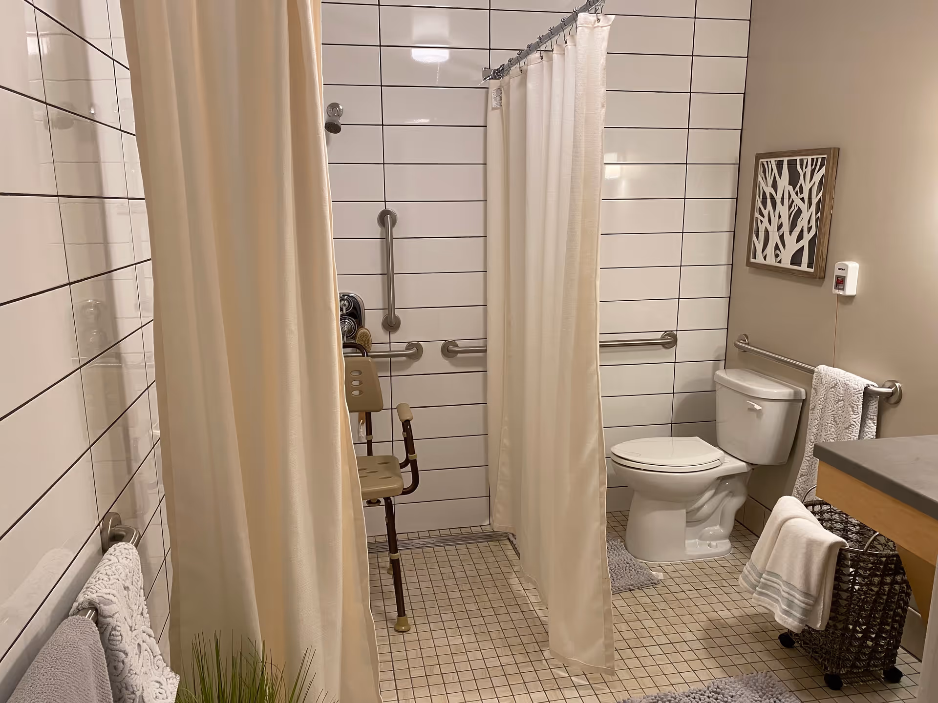 A bathroom with beige tiled floor and white tiled walls. There is a shower area with a beige shower curtain and a shower chair inside. The bathroom also has a white toilet, a towel rack with a white towel, a basket with a folded towel, and a wall decoration featuring a tree design. Safety grab bars are installed on the walls near the toilet and shower area.