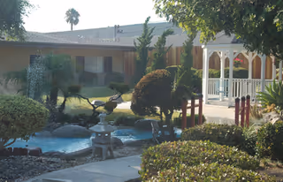 Outdoor garden area at Clara Baldwin Stocker Home featuring trimmed bushes, a small water feature with a bird statue, a white gazebo, and surrounding trees and plants.