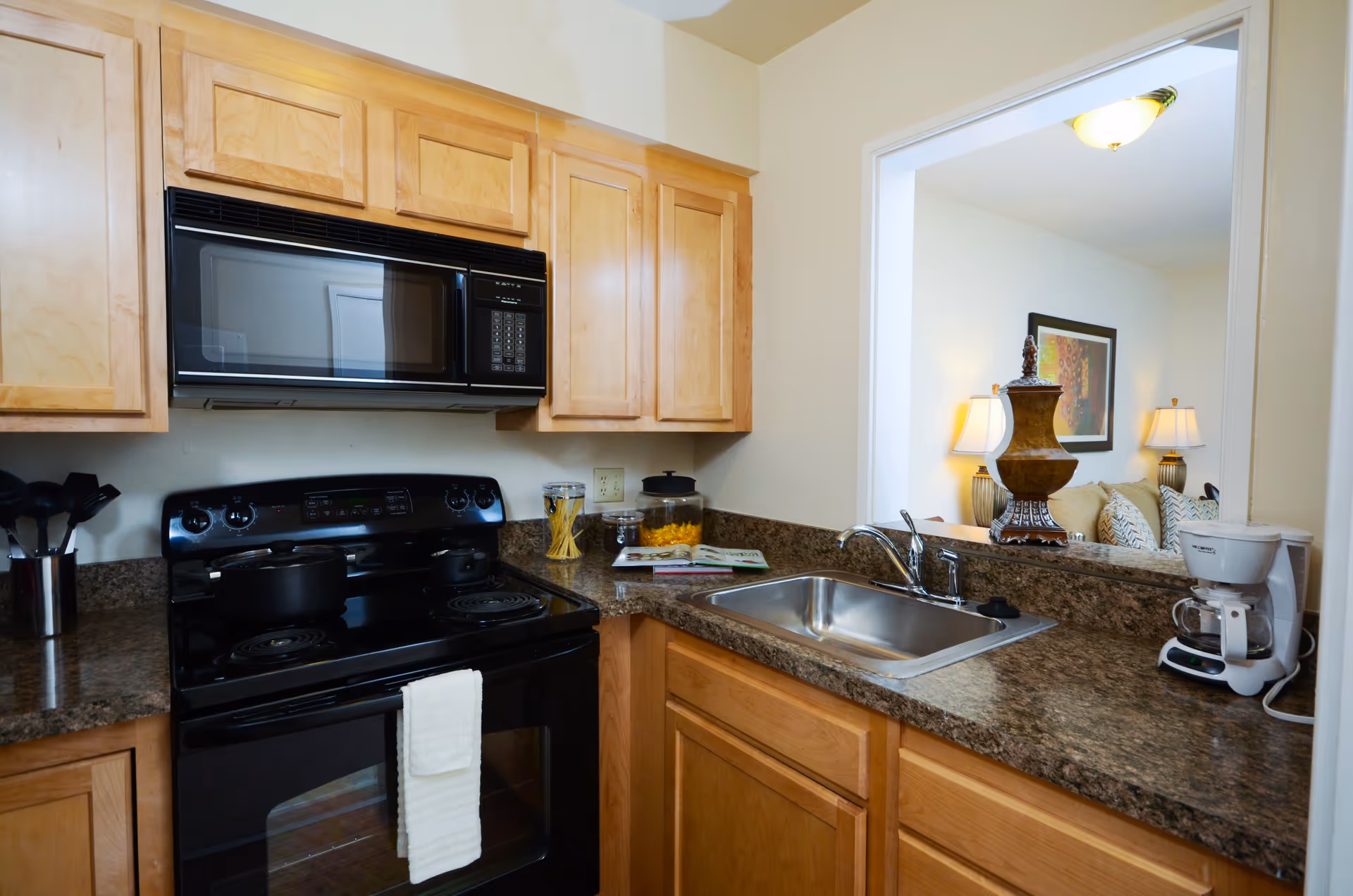 A compact kitchen with wooden cabinets, a black electric stove with a pot and pan on it, a microwave above the stove, a stainless steel sink, and a coffee maker on the countertop. Through a pass-through window, a living room area with a lamp, decorative vase, and framed artwork is visible.