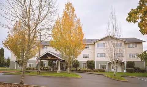 Front exterior view of a multi-story senior living building with fall trees and a driveway.