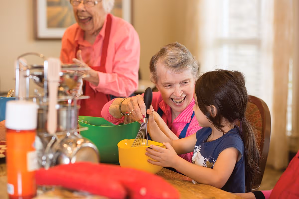 An elderly woman and a young girl are smiling and engaging in a cooking activity together. The elderly woman is helping the girl whisk ingredients in a yellow bowl, while another elderly woman in a pink shirt and red apron is laughing in the background. They are in a warm, well-lit kitchen setting with various kitchen utensils and bowls on the table.