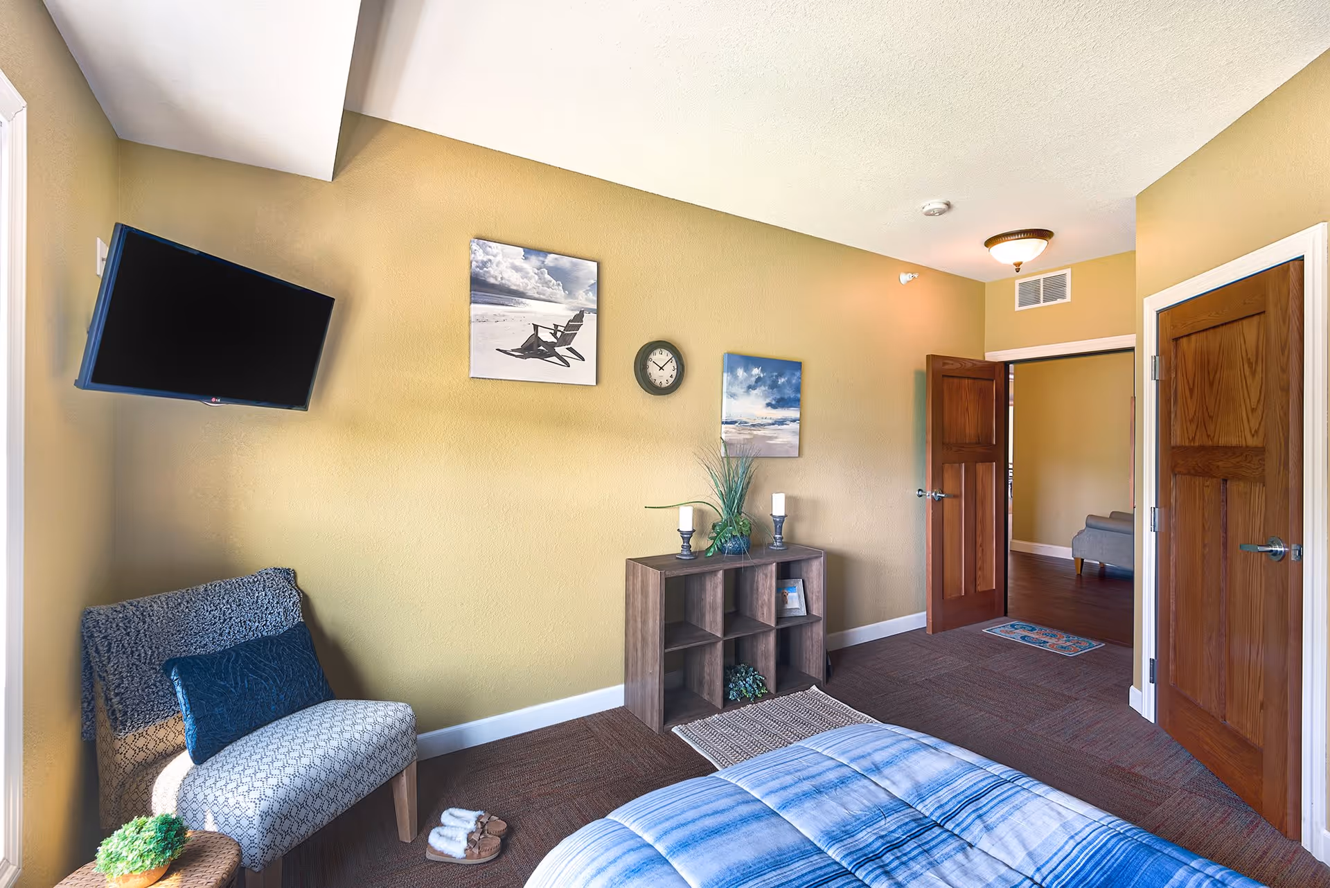 A cozy bedroom with a bed covered in a blue and white striped blanket. There is a patterned chair with a dark blue pillow next to a small round table with a plant on it. A flat-screen TV is mounted on the wall above the chair. The wall is painted yellow and decorated with two framed pictures and a round clock. A wooden shelf unit holds decorative items and candles. Two wooden doors are visible, one open leading to another room with a gray chair and wooden floor.