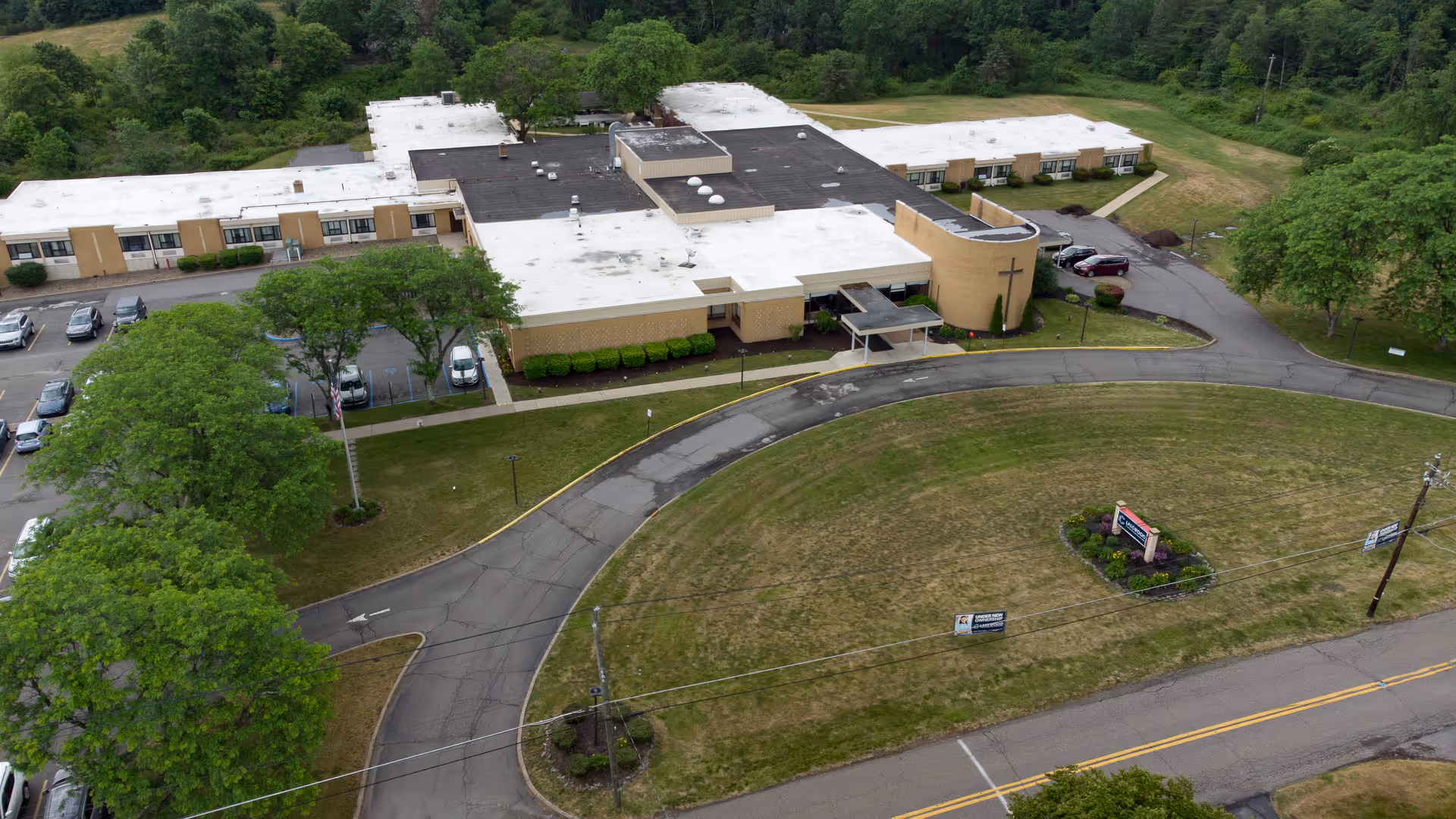 Aerial view of Lakewood Rehabilitation and Healthcare Center showing a large single-story building with a white and black roof, surrounded by green lawns, trees, and a curved driveway leading to the entrance. Several cars are parked in the parking lot adjacent to the building.