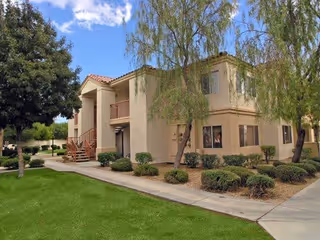 Exterior view of a two-story beige residential building with a red-tiled roof, surrounded by green bushes, trees, and a well-maintained lawn under a partly cloudy sky.