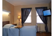 A small bedroom with a single bed covered in a light blue bedspread, a wooden tray table on the bed, a floor lamp beside the bed, two windows with dark curtains, and a wall-mounted television on the right side.