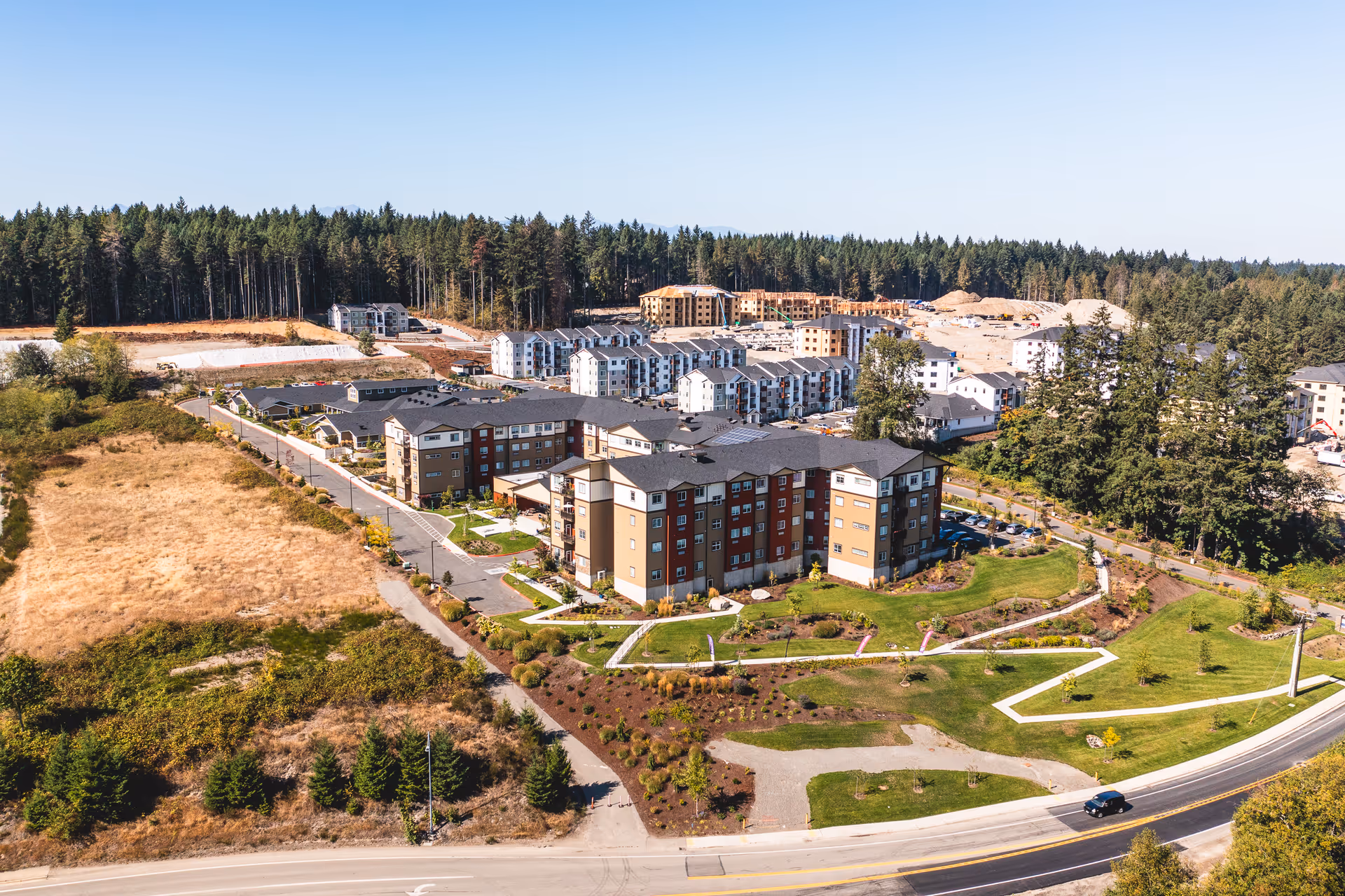 Aerial view of Fieldstone on Clear Creek facility showing multiple multi-story residential buildings surrounded by landscaped green areas, pathways, and a road in the foreground. The background includes a forested area and some construction activity.
