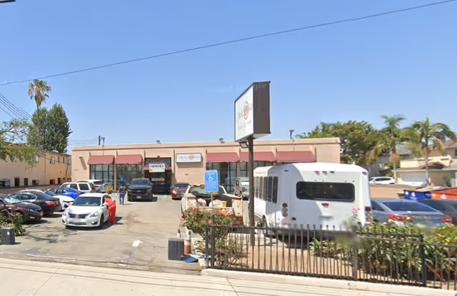 Exterior view of Coral Oaks Care Living facility showing a single-story building with a parking lot in front. Several cars and a white shuttle bus are parked. There is a signpost with the facility's name near the entrance. The sky is clear and blue.