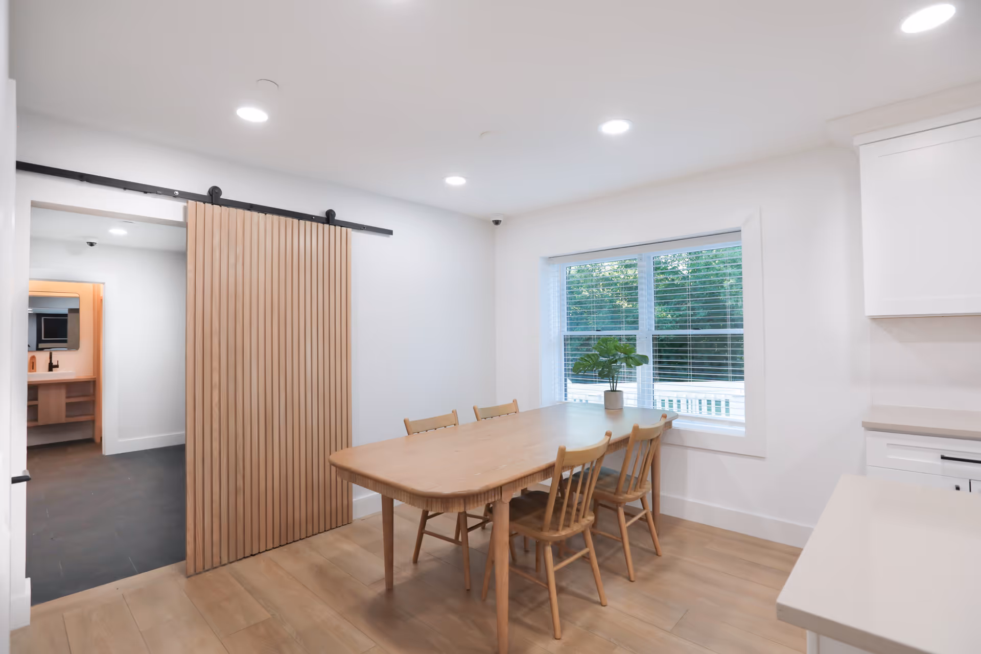 A bright dining area with a wooden table and four matching chairs. A small potted plant is placed on the table near a window with white blinds. The room features light wood flooring, white walls, and recessed ceiling lights. To the left, there is a sliding wooden door on a black metal track, partially open to reveal a hallway and another room.