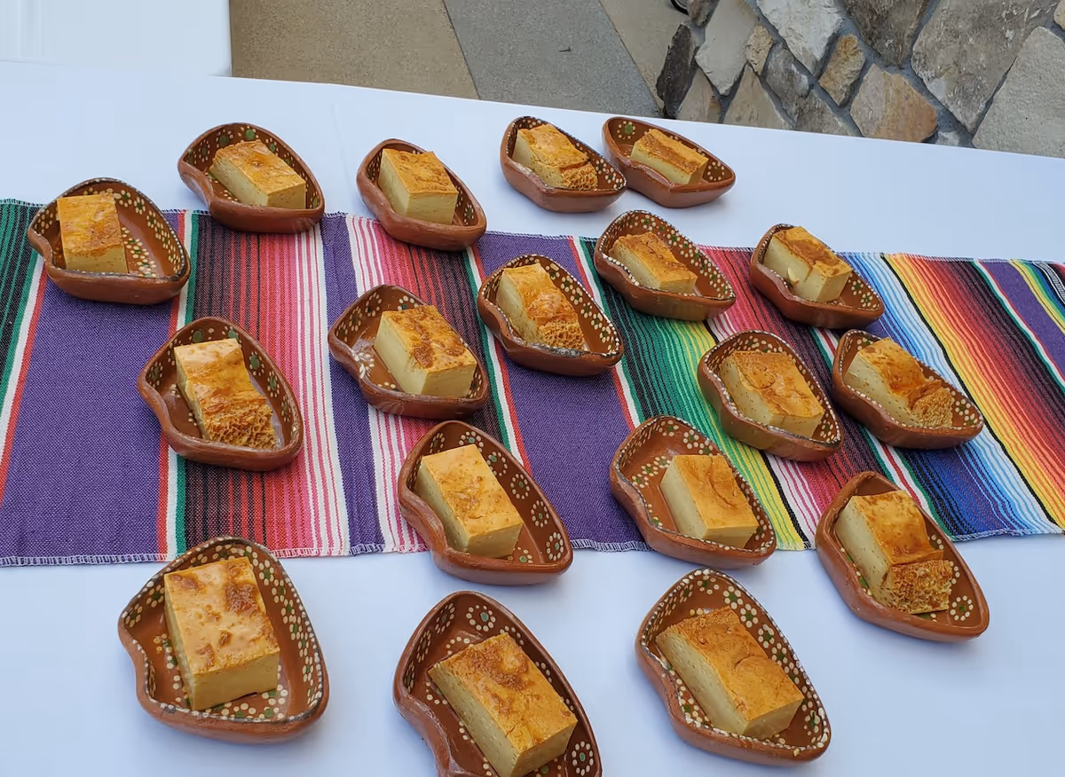 Multiple small brown ceramic dishes each containing a rectangular piece of baked dessert, arranged on a colorful striped table runner over a white tablecloth, with a stone wall and concrete floor in the background.