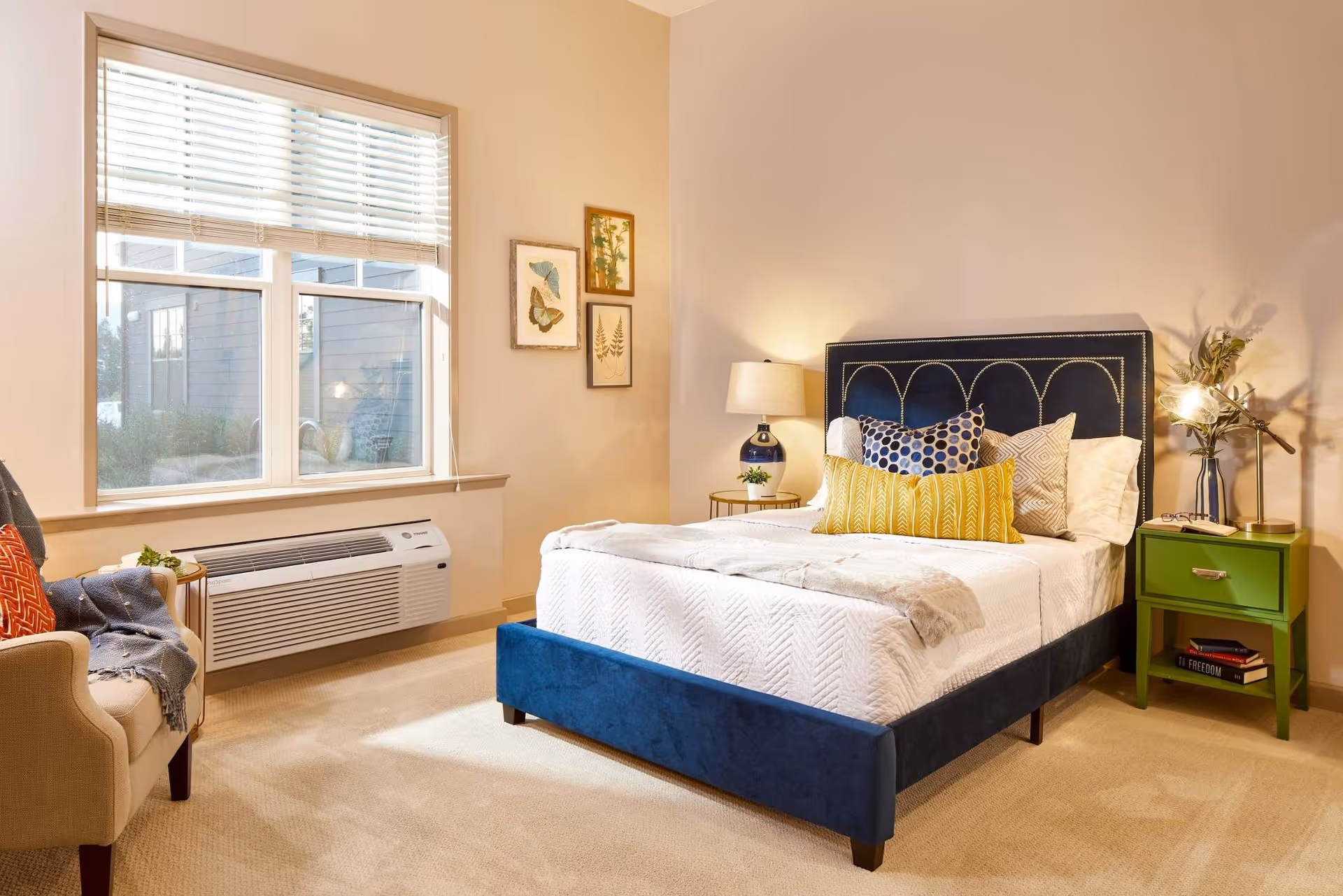A bright bedroom with a navy upholstered bed dressed in white linens, patterned pillows, a green nightstand, and an armchair by a large window.