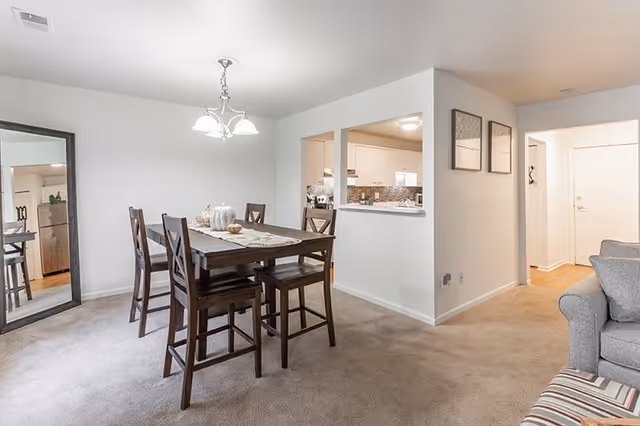 A dining area with a wooden table and six matching chairs on a carpeted floor. A chandelier with three lights hangs above the table. To the left, there is a large mirror leaning against the wall. The dining area opens into a kitchen with white cabinets and a tiled backsplash. On the right side, part of a gray sofa and a striped ottoman are visible, along with a hallway leading to a door. The walls are painted white, and two framed artworks hang on the wall near the hallway.