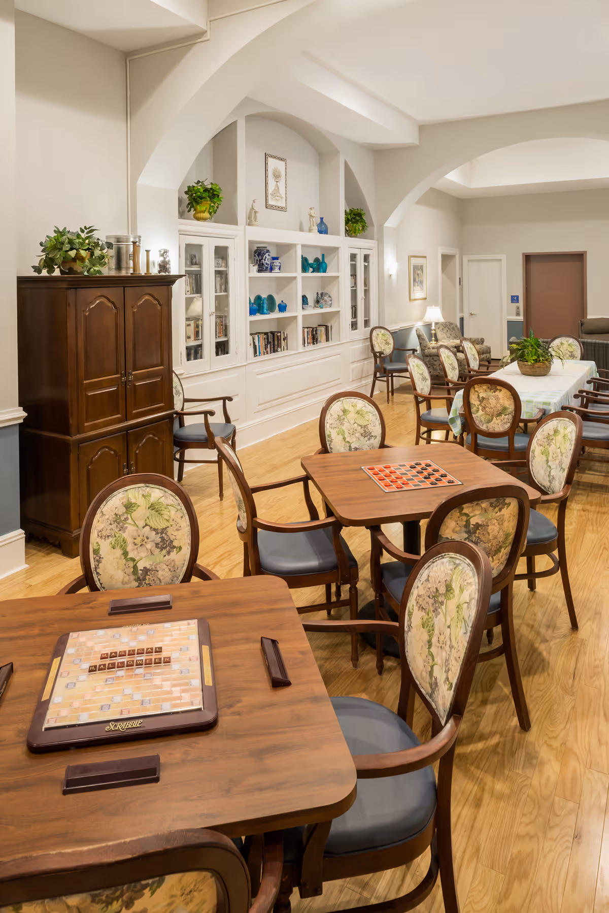 Bright common room with wooden game tables and floral-upholstered chairs, built-in shelving, and a long dining table in the background.