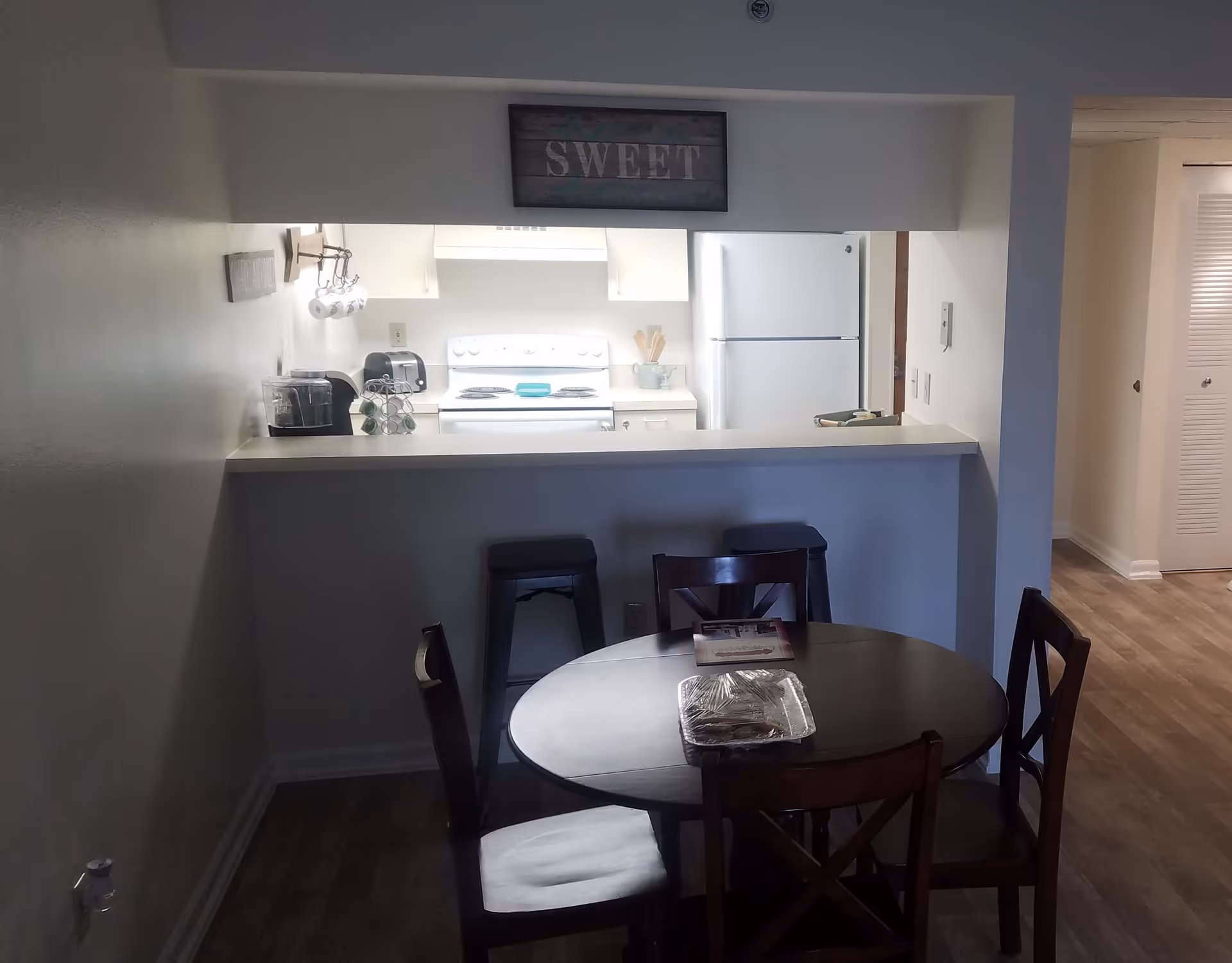 Round dining table with four chairs in front of a kitchen pass-through and two stools, with a 'SWEET' sign above the opening.