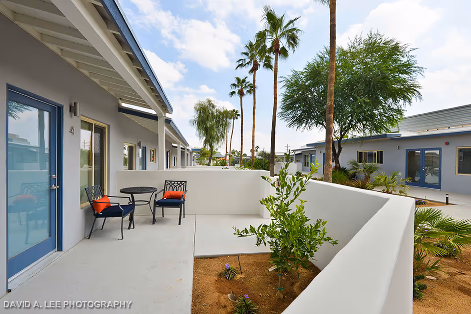 Outdoor courtyard patio of a single-story assisted living building with a small table and two chairs, low white walls, potted plants and palm trees in the background.