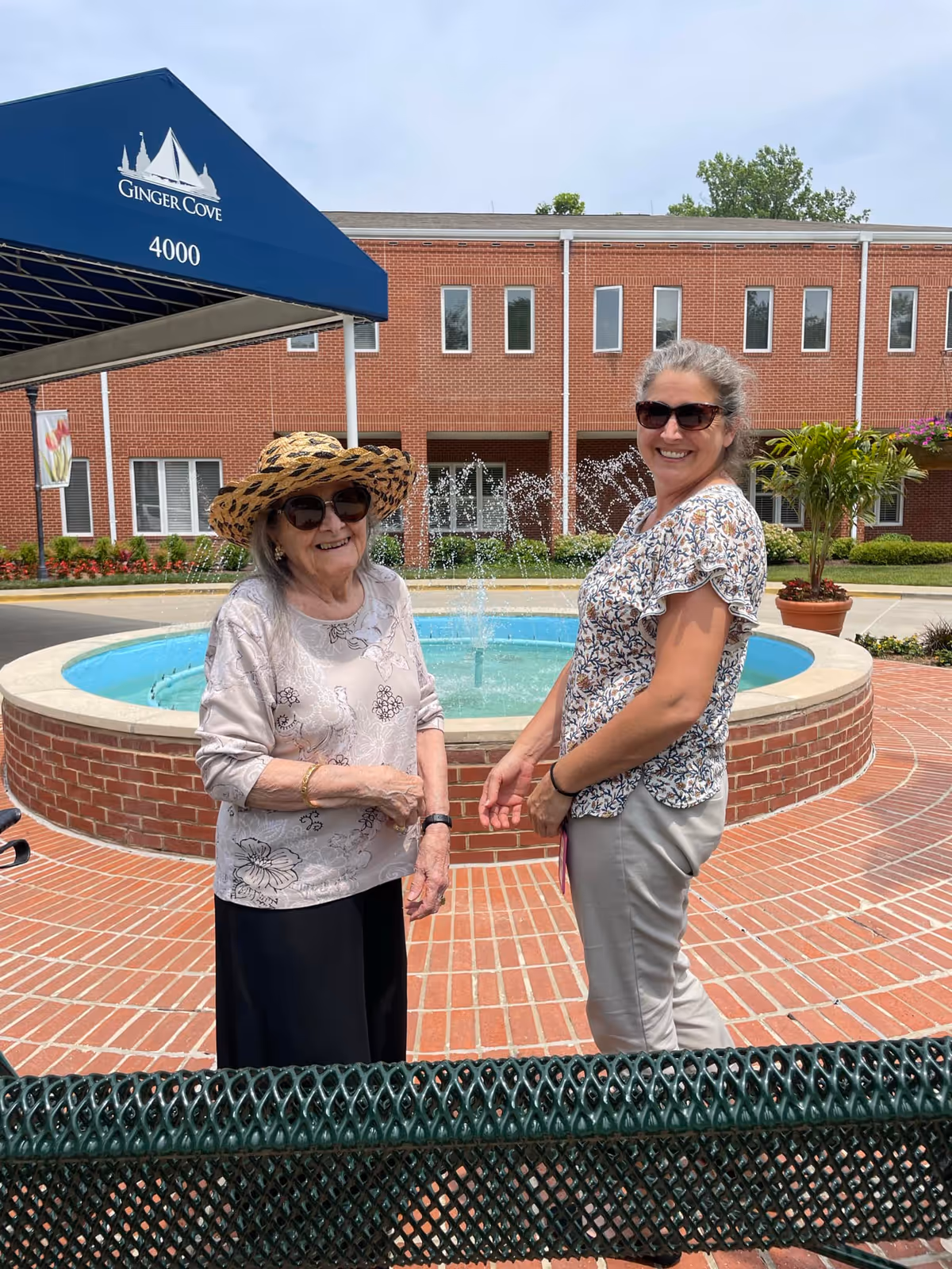 Two women wearing sunglasses stand in front of a circular fountain outside a brick building with a blue 'Ginger Cove' awning.