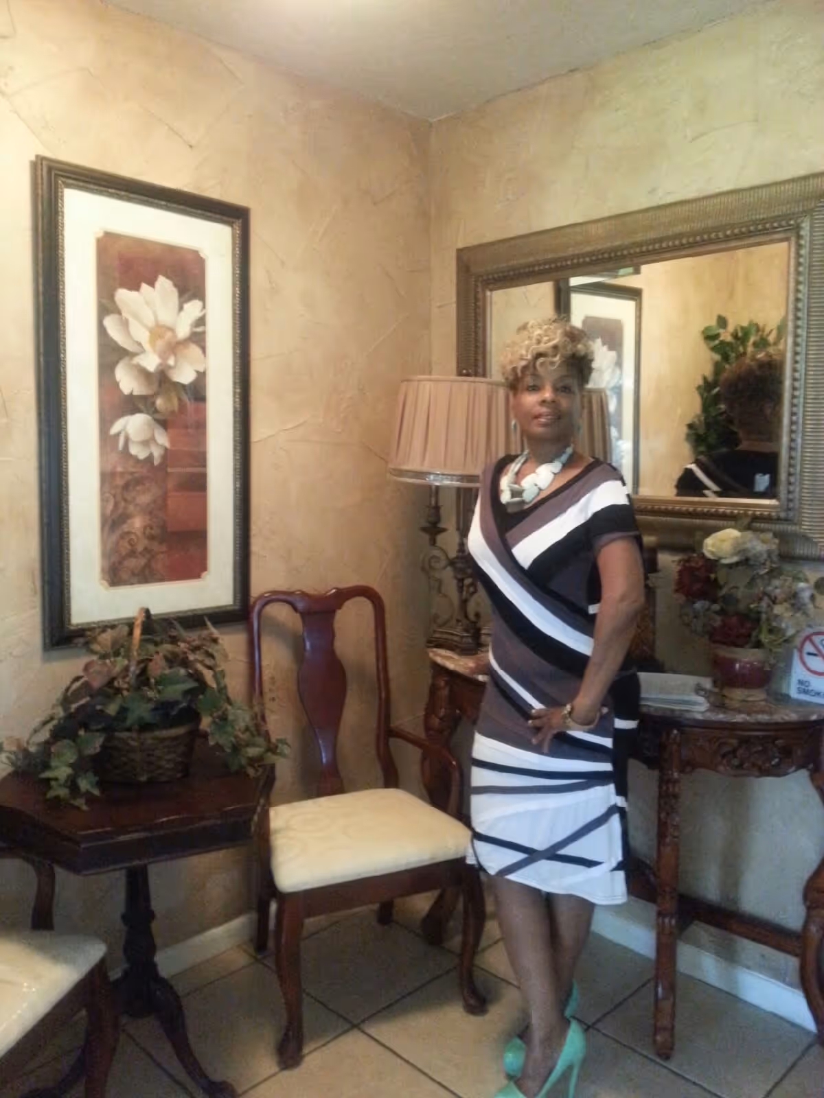 A woman standing in a warmly decorated room with beige textured walls. She is wearing a black, white, and gray striped dress with turquoise high heels and a chunky necklace. Behind her is a large ornate mirror, a table with a lamp, and a floral arrangement. To her left are two wooden chairs with cream cushions and a small table with a basket of greenery. A framed floral artwork hangs on the wall.