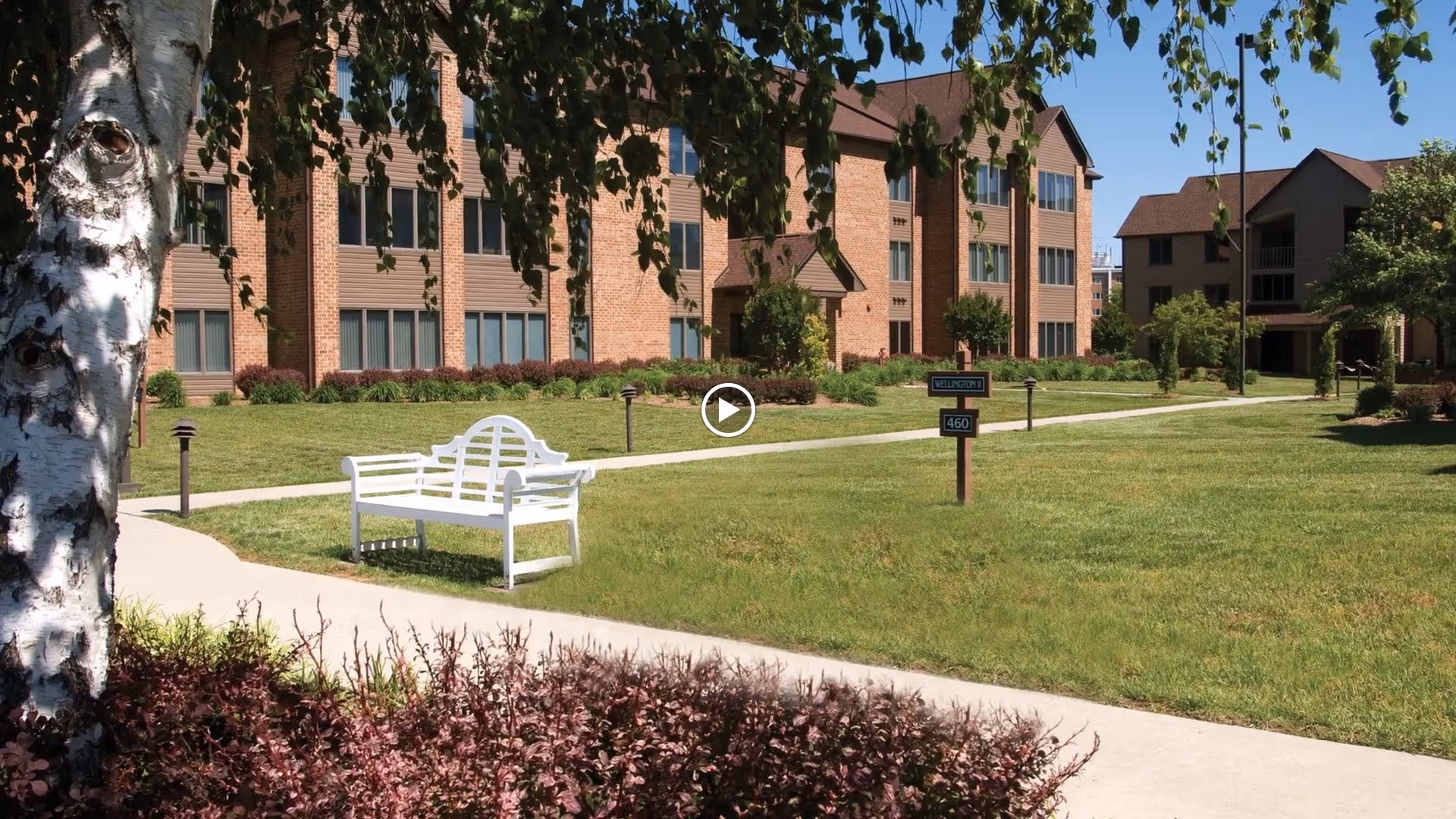 Outdoor view of a senior living facility with a well-maintained lawn, a white bench on a concrete pathway, trees, and multi-story brick buildings in the background under a clear blue sky.